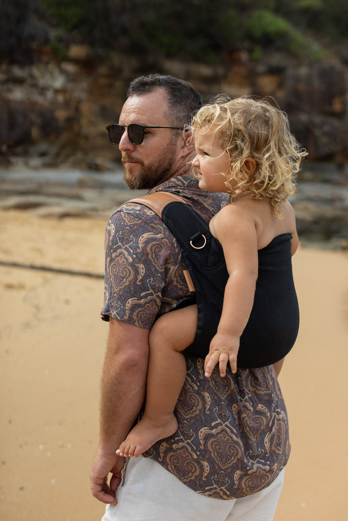 Dad carrying toddler in black Mamatoto Nomad ergonomic baby carrier at the beach — hip-healthy toddler carrier.