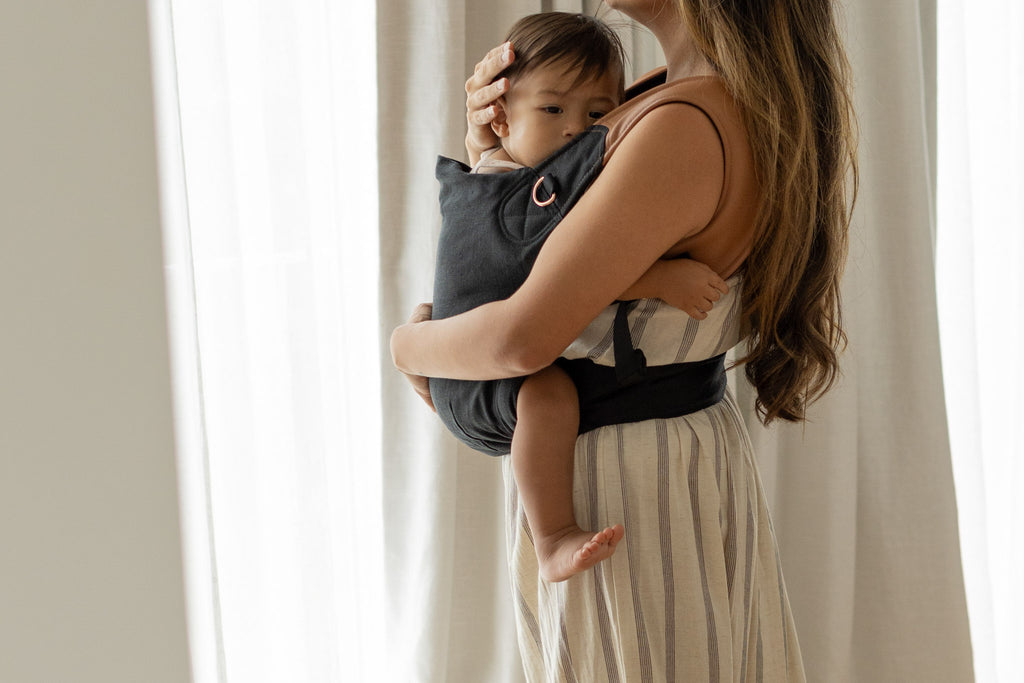 Baby cuddled in black Mamatoto Nest ergonomic baby carrier indoors — soft, supportive newborn carrier for mums.